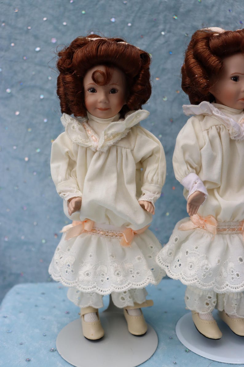 May include: Two porcelain dolls dressed in cream-colored outfits with lace trim and peach-colored bows. The dolls have brown curly hair and are standing on a light blue surface. The dolls are wearing matching outfits, including dresses and pants.