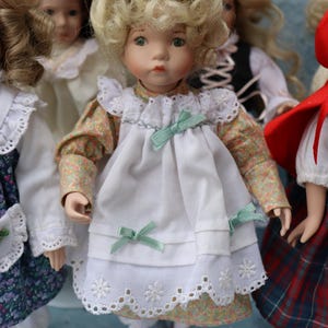 May include: A porcelain doll with blonde curly hair, wearing a white dress with a floral print underdress and mint green bows. The doll has red shoes and is surrounded by other dolls.