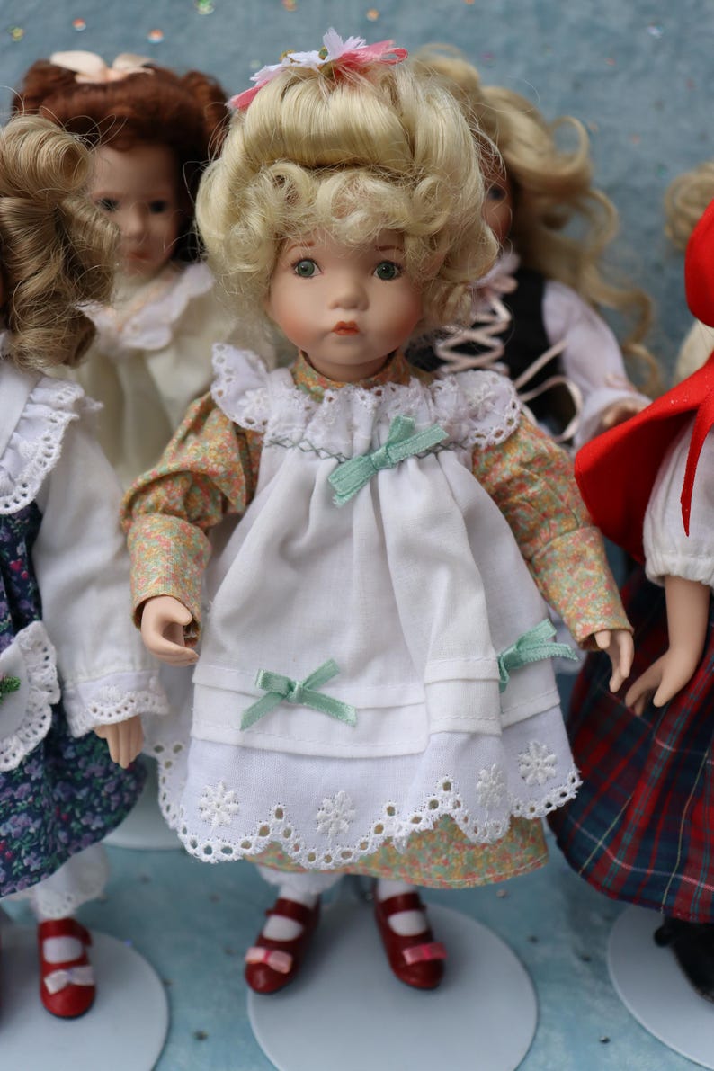 May include: A porcelain doll with blonde curly hair, wearing a white dress with a floral print underdress and mint green bows. The doll has red shoes and is surrounded by other dolls.
