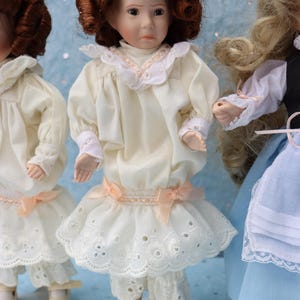 May include: Three porcelain dolls are displayed, each with detailed features. The central doll wears a cream-colored dress with a lace-trimmed skirt and sleeves, accented by a peach bow. The dolls have brown curly hair and are on white stands.