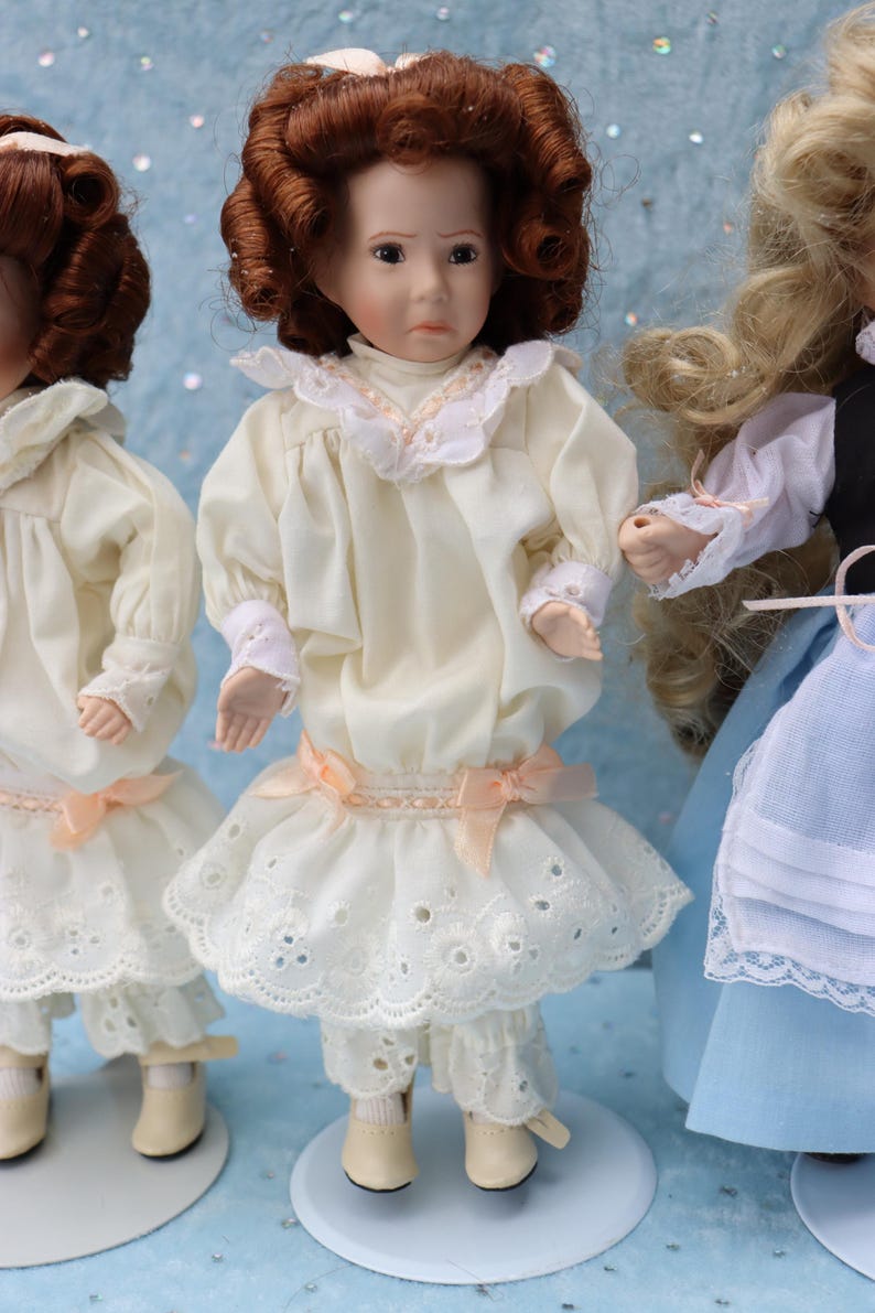 May include: Three porcelain dolls are displayed, each with detailed features. The central doll wears a cream-colored dress with a lace-trimmed skirt and sleeves, accented by a peach bow. The dolls have brown curly hair and are on white stands.