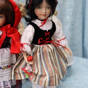 May include: A porcelain doll dressed in a Snow White costume. The doll has black hair, blue eyes, and is wearing a white blouse, a black vest, and a striped skirt. She holds a red apple and a small bouquet.