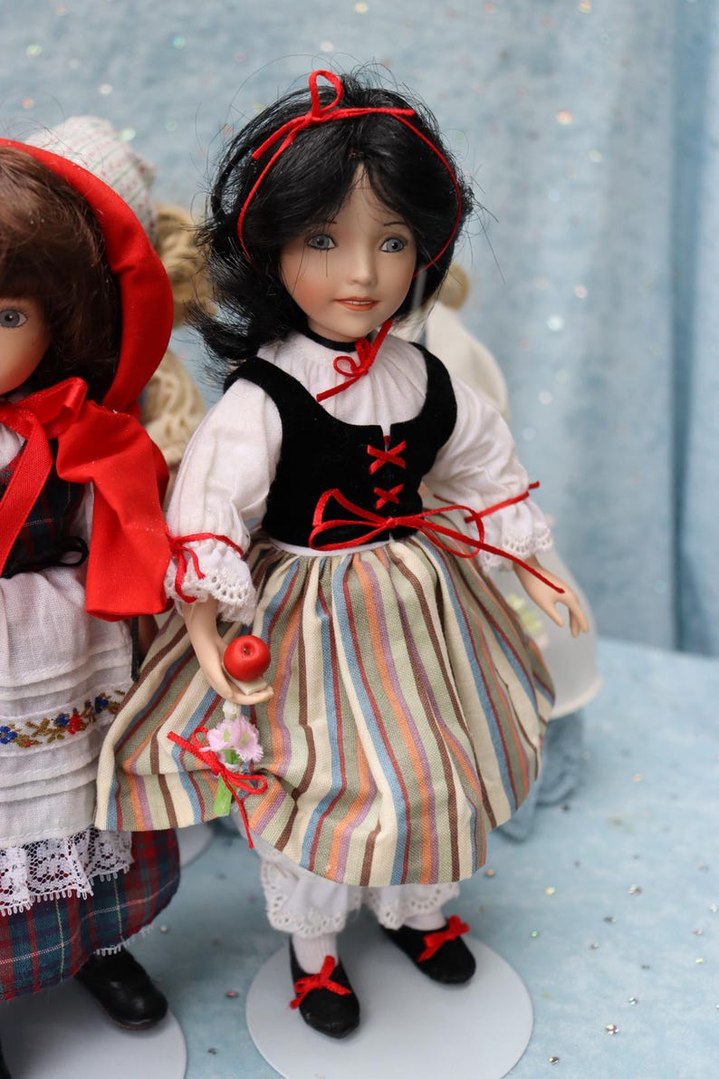 May include: A porcelain doll dressed in a Snow White costume. The doll has black hair, blue eyes, and is wearing a white blouse, a black vest, and a striped skirt. She holds a red apple and a small bouquet.