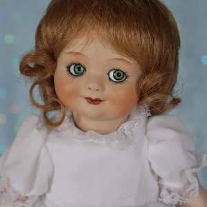 Könnte beinhalten: Eine Porzellanpuppe mit rotbraunem Haar, das in weichen Locken gestylt ist. Die Puppe hat grüne Augen, rosige Wangen und ein zartes Lächeln. Sie trägt ein weißes Kleid mit Spitzensaum am Ausschnitt und an den Ärmeln. Der Hintergrund ist ein sanftes Blau mit Bokeh.