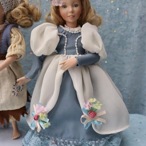 May include: A doll in a blue and white dress with a pearl necklace and blonde hair. The dress has white puffy sleeves and a lace hem. Small floral accents adorn the dress. Another doll is partially visible on the left.