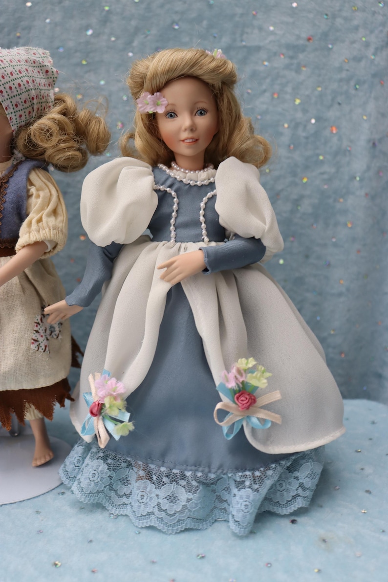 May include: A doll in a blue and white dress with a pearl necklace and blonde hair. The dress has white puffy sleeves and a lace hem. Small floral accents adorn the dress. Another doll is partially visible on the left.