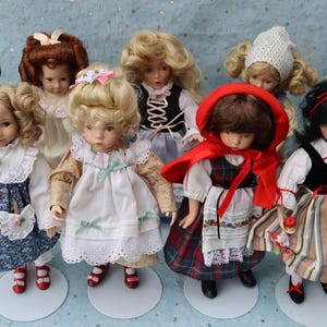May include: A collection of vintage porcelain dolls, each with unique hairstyles and outfits. The dolls are dressed in various styles, including floral dresses, a red cape, and a black vest. They stand on small, round white bases.