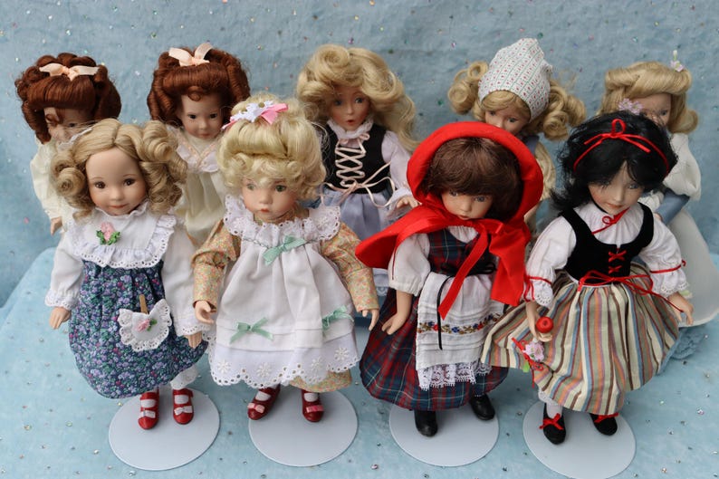May include: A collection of vintage porcelain dolls, each with unique hairstyles and outfits. The dolls are dressed in various styles, including floral dresses, a red cape, and a black vest. They stand on small, round white bases.