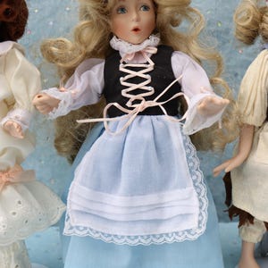 May include: A porcelain doll with blonde hair, blue eyes, and a black vest over a white blouse. The doll wears a light blue skirt and apron with lace trim. The doll is part of a collection of porcelain dolls.