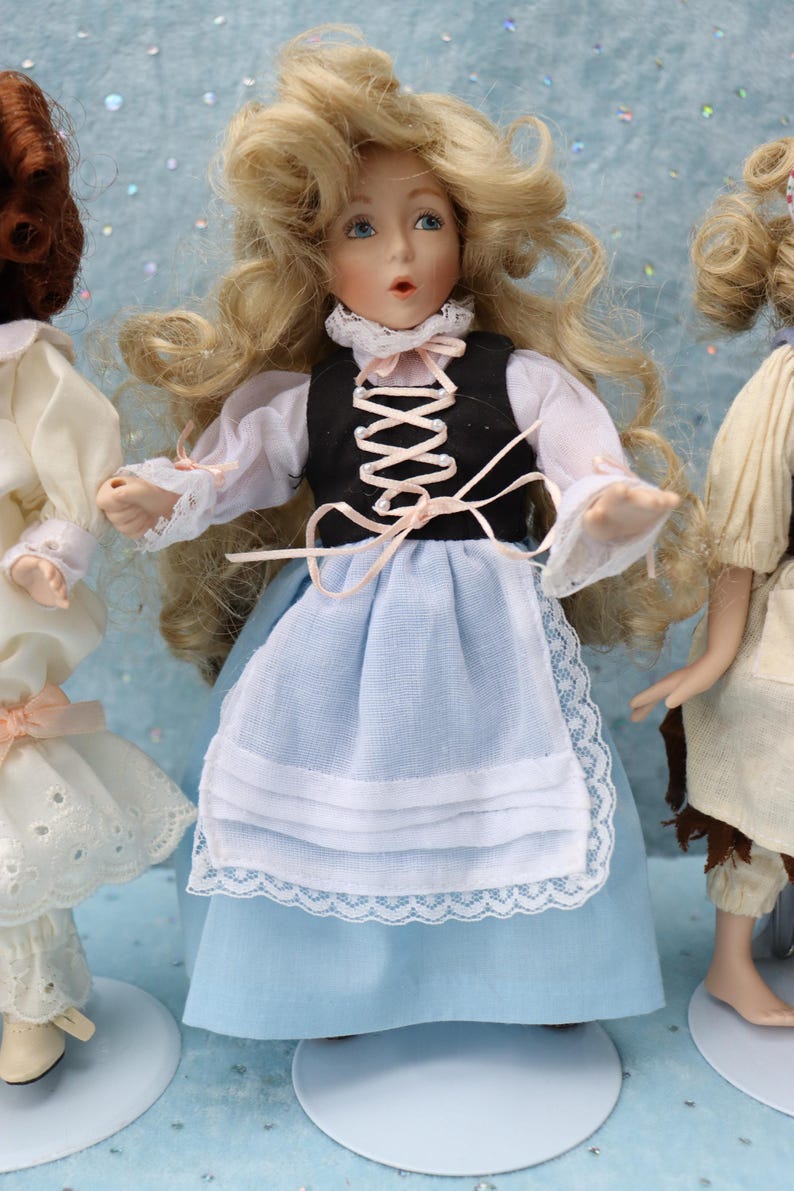 May include: A porcelain doll with blonde hair, blue eyes, and a black vest over a white blouse. The doll wears a light blue skirt and apron with lace trim. The doll is part of a collection of porcelain dolls.
