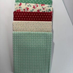 May include: A stack of five folded fabric squares featuring various patterns. The top piece is green with small white crosses, followed by a cream fabric with pink and teal flowers, a deep red piece with white dots, a cream fabric with tiny floral sprigs, and a teal piece with a geometric cross pattern.