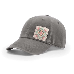 May include: A charcoal gray baseball cap featuring a square patch on the front panel. The patch displays a geometric star pattern in shades of pink, green, and cream, with a small checkered design integrated into the colorful star motif.