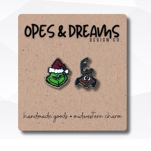 May include: Two Christmas-themed enamel pins on a brown card. One pin features a green character with a Santa hat, the other a brown character. The card reads "OPES & DREAMS DESIGN CO." and "handmade goods • midwestern charm."