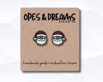 Puede incluir: Pendientes con la cara de Papá Noel sobre una tarjeta marrón. Los pendientes tienen un gorro rojo, barba blanca y una cara sonriente. La tarjeta tiene el texto "OPES & DREAMS DESIGN CO." y "handmade goods • midwestern charm."