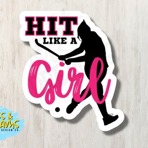 以下が含まれることがあります： ピンクと黒のステッカー。ホッケーのスティックを持った人のシルエットと「HIT LIKE A GIRL」の文字が書かれています。
