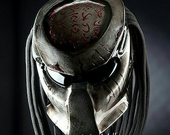 Può includere: Un casco da moto argento e nero con un design ispirato al Predator. Il casco ha una visiera trasparente e un cinturino per il mento nero intrecciato. La parte superiore del casco presenta un design circolare rosso con segni intricati.