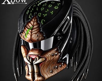 May include: A black motorcycle helmet featuring a textured, tan-colored creature-like face mask with prominent fangs, a dark visor, and long black braided hair-like strands. The top of the helmet displays glowing green symbols and the text XBow.