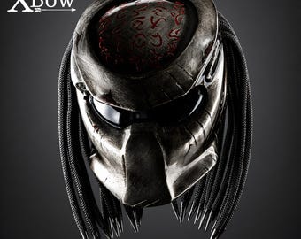May include: A full-face helmet with a weathered metallic silver finish, featuring a dark visor and a top section adorned with red etched symbols. The helmet includes long, black, textured dreadlock-style strands on both sides and the text XBow.