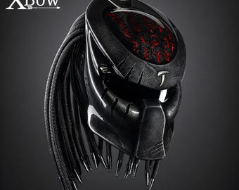 May include: A black motorcycle helmet featuring a textured finish, a wide visor opening, and a series of long, black braided cords extending from the back. The top section displays a recessed panel with intricate red glowing symbols and the text XBow.