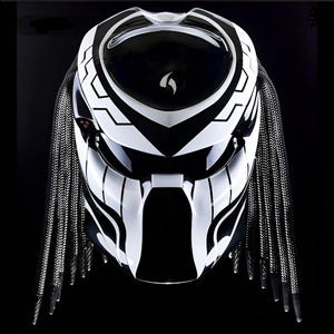 Puede incluir: Un casco de moto blanco y negro con un diseño inspirado en Predator. El casco tiene una visera negra y un patrón blanco y negro en la parte delantera. El casco tiene una correa trenzada blanca y negra que cuelga hacia abajo en la parte posterior.