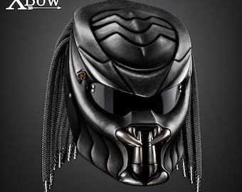 May include: A matte black full-face helmet featuring a textured, sculpted exterior with a dark tinted visor, a prominent jaw guard with metallic fangs, and long black braided cords hanging from the sides. The text XBow is visible in white.