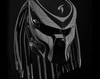 Può includere: Un casco da moto nero e bianco con un design ispirato a Predator. Il casco ha una visiera nera e una lunga treccia nera.