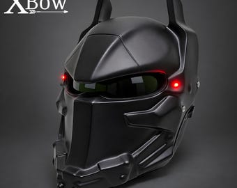May include: A matte black full-face helmet featuring a stylized bat-like silhouette with two pointed ears on top. The helmet includes a dark visor, a prominent central nose guard, and two small red LED lights glowing on the sides. The text XBow is visible.