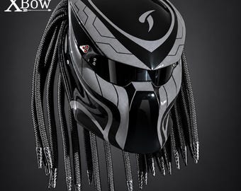 May include: A black and silver motorcycle helmet featuring the text XBow. The helmet has a glossy finish with geometric patterns, a dark visor, and numerous long, braided black cords hanging from the sides and back, each tipped with silver metal.