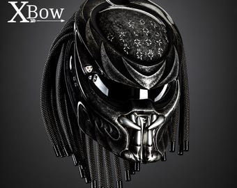 May include: A black motorcycle helmet featuring a textured, dark metallic finish with a Predator-inspired design. The helmet includes a visor, a sculpted faceplate with metallic fangs, and numerous black braided cords hanging from the sides. Text XBow.