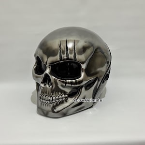 Può includere: Un casco a forma di teschio argentato con un design scheletrico dettagliato. Il casco presenta un design integrale con orbite e mascella. Il testo "XbowHelmets" è visibile sul lato.