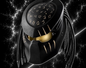 May include: A matte black full-face helmet featuring a gold-tinted visor, gold-colored claw mark accents on the forehead and cheek, and gold-colored alien symbols on the top. The helmet includes long, black braided cords hanging from the sides.