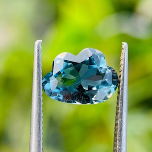 May include: A faceted, cloud-shaped gemstone with a deep blue color and a high-gloss, reflective surface. The stone is held by two metallic, serrated-edge tweezers, showcasing its intricate, multi-faceted cut and translucent quality.
