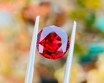 May include: A faceted, round, deep red gemstone held by silver tweezers. The gem has a brilliant cut, reflecting light. The background is blurred, with hints of green and yellow.