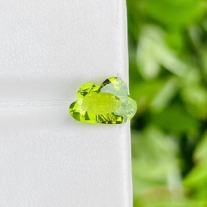 Pode incluir: Uma pedra preciosa facetada em forma de nuvem, em um tom verde claro vibrante. A pedra preciosa está colocada sobre um fundo branco, com um fundo desfocado de folhagem verde. O corte da pedra preciosa revela facetas internas.