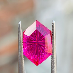 May include: A faceted, hexagonal gemstone in a vibrant magenta hue. The gem is held by silver tweezers, showcasing its intricate internal patterns. The cut of the stone creates a starburst effect, reflecting light. The gemstone is a striking example of fine jewelry.