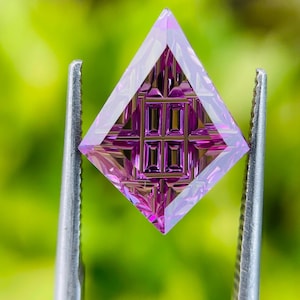 May include: A kite-shaped gemstone with a vibrant magenta and purple hue, featuring a complex, precision-cut geometric grid pattern on its surface. The stone displays sharp, symmetrical facets that create deep internal reflections and light play.