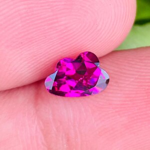 Peut inclure: Une pierre précieuse en forme de cœur, taillée à facettes, de couleur magenta vif. La taille de la gemme révèle de multiples facettes, créant un effet scintillant. La pierre est tenue entre les doigts, sur fond rose.