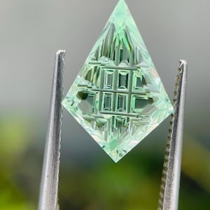 May include: A kite-shaped, light mint green gemstone featuring a complex, geometric precision-cut pattern on its surface. The stone is held by silver-toned metal tweezers, showcasing its translucent quality and sharp, angular facets.