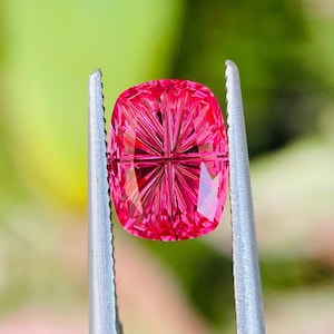 Può includere: Una gemma rosa brillante, taglio a cuscino, tenuta da una pinzetta. La gemma presenta sfaccettature complesse e un motivo interno. Lo sfondo verde sfocato mette in risalto il colore e la brillantezza della pietra.