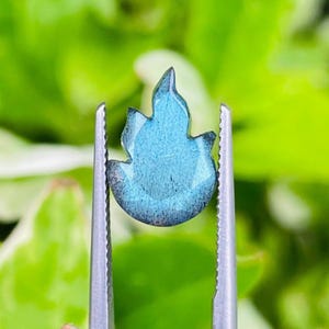 Könnte beinhalten: Ein blauer, flammenförmiger Edelstein wird von einer silbernen Pinzette vor einem verschwommenen grünen Hintergrund gehalten. Der Edelstein hat eine facettierte Oberfläche und einen dunklen Rand. Die Pinzette ist scharf, was die Details des Edelsteins hervorhebt.