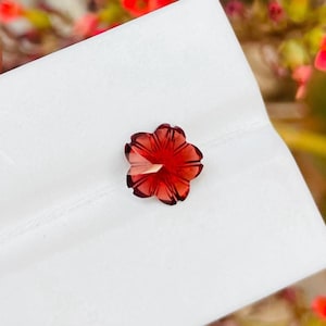 Könnte beinhalten: Ein facettierter, roter Edelstein in Form einer Blume. Der Edelstein hat eine transluzente Qualität mit dunkleren roten Rändern und einem helleren roten Zentrum. Die Blume liegt auf einer weißen Oberfläche.