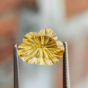 May include: A cloud-shaped, golden-yellow gemstone is held by silver tweezers. The gem is intricately faceted with radiating lines, creating a sunburst pattern. The blurred background accentuates the gemstone's details. The gem is measured in centimeters.
