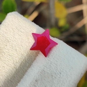 Puede incluir: Una gema facetada en forma de estrella con un degradado de rojo a rosa. La gema se presenta sobre un fondo blanco texturizado, resaltando su diseño geométrico y variaciones de color. La forma de estrella está bien definida.