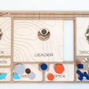 May include: Wooden game organiser with compartments labelled "DECK", "DREADS", "WATER", "SOL", "SPICE", "TOOLS", and "DISCARD". Includes a "LEADER" card and various game pieces in blue, orange, and grey. Features engraved geometric designs.