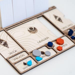 Puede incluir: Organizador de juegos de mesa de madera con ranuras para cartas y varias piezas de juego. El organizador tiene secciones etiquetadas como "Deck", "Dreads", "Water", "Leader", "Spice" y "Discard". Incluye piezas de juego azules, grises y naranjas.