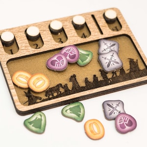 Puede incluir: Organizador de juegos de madera con ranuras para fichas y marcadores de jugador. El organizador tiene un interior marrón con una silueta de paisaje. Incluye varias fichas de juego en colores como morado, verde y naranja.