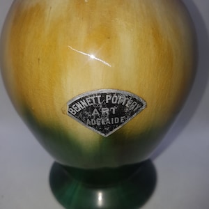 May include: A green and yellow ceramic vase with a round body and a flared base. The vase has a black and white label that reads "Bennett Pottery Art Adelaide".