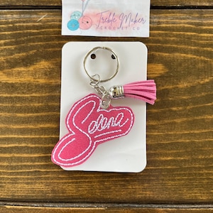 May include: A pink keychain with the name "Selena" embroidered in white. The keychain has a silver ring and a pink tassel. The keychain is attached to a white card with the text "Treble Maker Crochet Co."