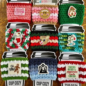 May include: A collection of handmade crochet cup cozies in various colors and designs. Some feature holiday-themed appliqués like a gingerbread man, reindeer, and gnome. Others have text patches that read "Candy Cane Cutie" and "Oh Snap".