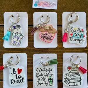 May include: A collection of embroidered keychains with various designs and phrases. The keychains feature phrases like "Reading is my therapy," "I like my books spicy & my coffee icy," and "Cowboy Romance Lover." Each keychain has a tassel.